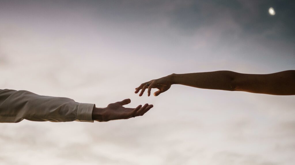 Two hands reaching towards each other against a soft twilight sky symbolizing connection and love.